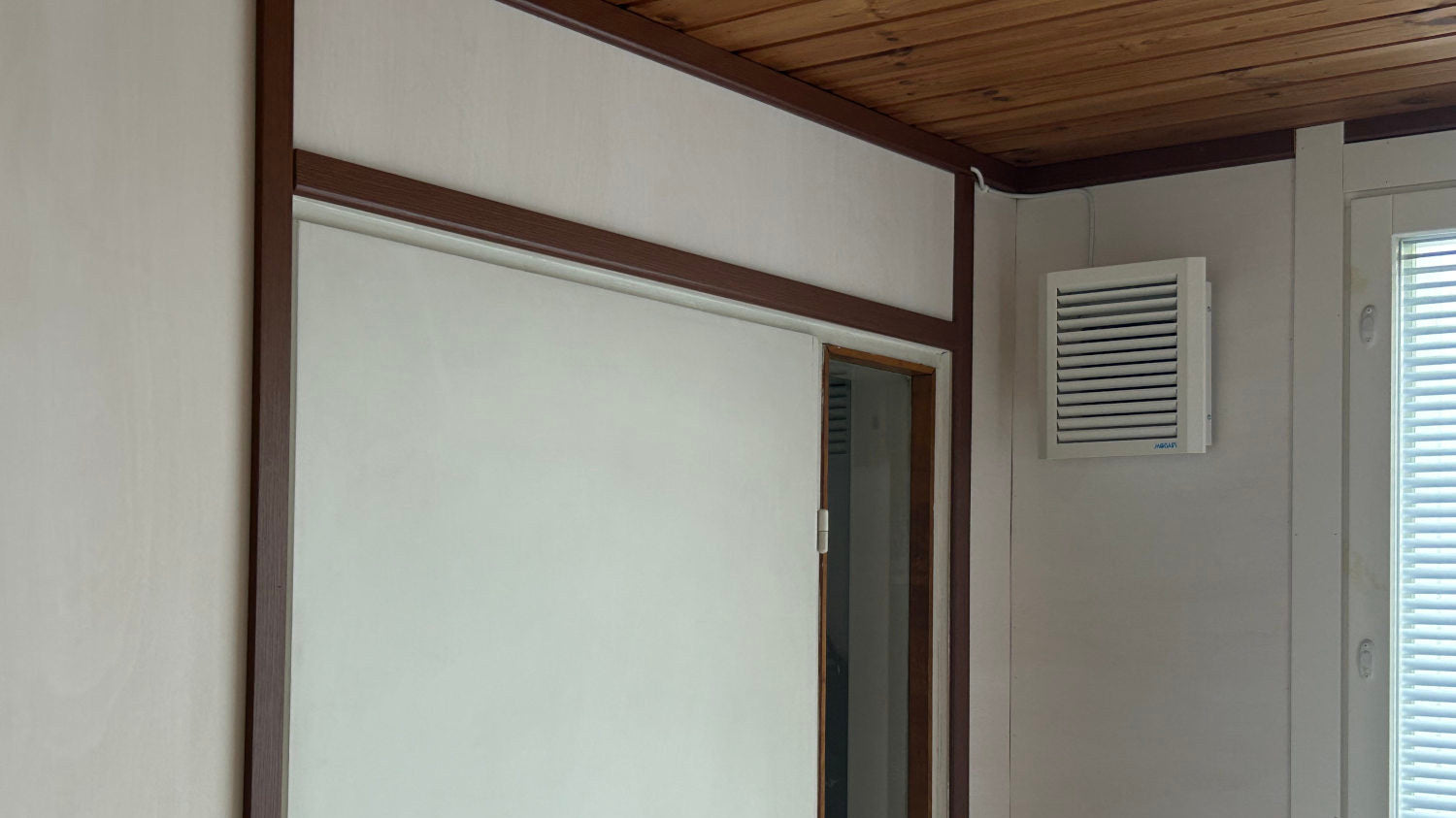 Room interior with a white door, wooden ceiling, and wall-mounted Mobair M fresh air valve.