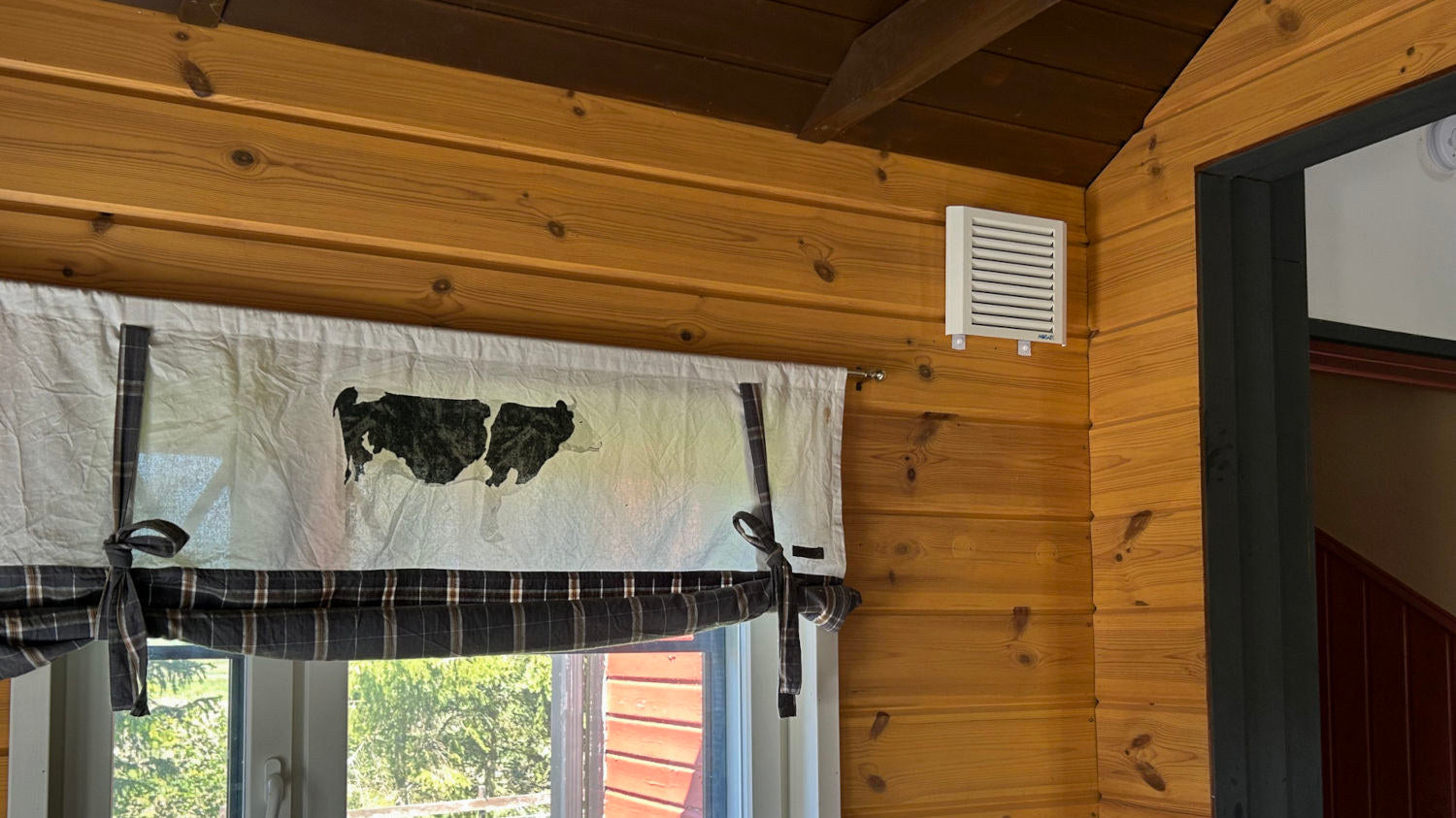Mobair Out exhaust fan mounted on the wall next to a window with curtain featuring cow print in a wooden cabin setting.