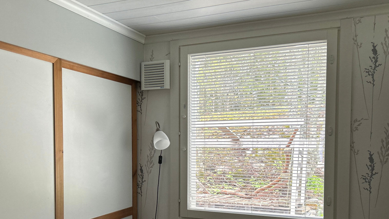 Room interior with a window with blinds, Mobair S fresh air valve mounted on a wall, and a light fixture.